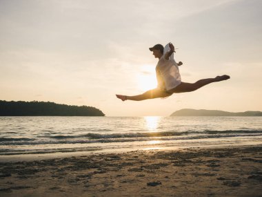 Gün batımında Malezya, Langkawi 'de bir sahilde canlı bir atlayış gerçekleştiren bir insan, dingin okyanus zeminine karşı neşe ve özgürlüğü ifade ediyor. Bir coşku anı.