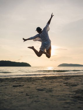 Gün batımında Langkawi, Malezya 'da bir plajda neşeyle sıçrayan bir insan, huzurlu bir okyanus zeminine karşı özgürlük ve mutluluğu somutlaştırıyor - bir yaşam kutlaması.