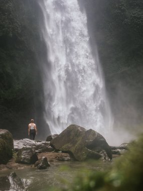 Yalnız bir gezgin güçlü bir şelalenin önünde duruyor Bali 'nin ormanındaki yemyeşil bir kayalıktan aşağı yuvarlanıyor, etrafını çevreleyen sis ve canlı yeşillik - Tropik Cennet' te Huzur ve macera