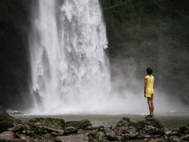 Sarı kıyafetli bir adam, güçlü bir şelale tarafından büyülenmiş ve Bali ormanının yemyeşil yeşiliyle çevrili bir havuza atlayan kayalık bir arazide duruyor.