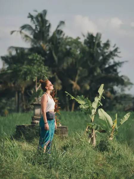 Bir kadın yemyeşil çayırlarda duruyor, ılık güneş ışığının tadını çıkarıyor ve Bali 'nin doğal ortamının barışçıl atmosferini kucaklıyor - Doğanın kucaklamasındaki farkındalık ve sükunet