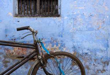 A classic bicycle with a brown leather seat and handlebars rests against a weathered blue wall. The bicycle has cream colored tires and a silver fram