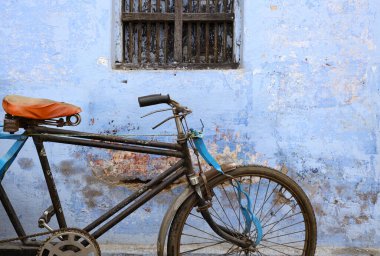 A classic bicycle with a brown leather seat and handlebars rests against a weathered blue wall. The bicycle has cream colored tires and a silver fram