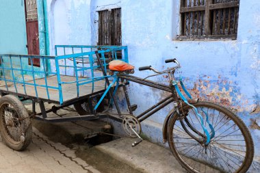 blue tricycle with a red basket attached to the back wheel stands parked on the side of a quiet residential street