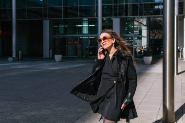 Yüzünde gülümseme olan yönetici kız işten sonra telefonla konuşuyor, arka planda ofis binaları var..