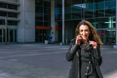 Şüpheli bir ifadesi olan yönetici kız kameraya bakıp telefonda konuşurken güneş gözlüğünü ısırıyor. Arka planda iş binası olan ofis dışında..
