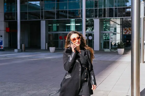 Female executive walks out of offices while talking on the phone and smiling, with office tower in background