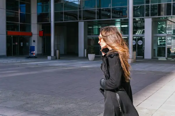 Profile view of executive girl with a negative expression on his face talking on the phone after work, with office buildings in the background.