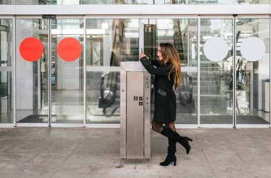 Bir iş merkezinin girişinde cep telefonuyla gülümseyen ve daktilo kullanan yönetici bir kız. Yürüyen merdivenler ve arka planda otomatik kapı var.