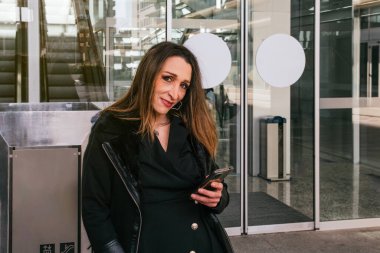 Akıllı telefonunu kullanan yönetici kız bir iş binasının girişinde kameraya gülümseyerek bakıyor.