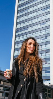 Yönetici kızın iş molasında kahve içtiği orta boy bir fotoğraf. Ofis gökdelenleri arka planda