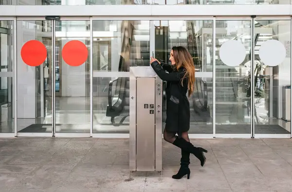 Bir iş merkezinin girişinde cep telefonuyla gülümseyen ve daktilo kullanan yönetici bir kız. Yürüyen merdivenler ve arka planda otomatik kapı var.