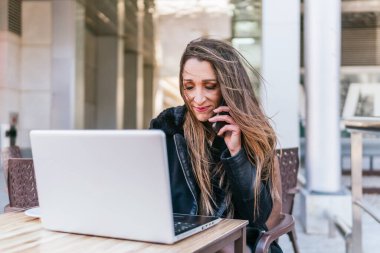 Yönetici kız, ofisinin yakınındaki bir barın terasında bilgisayarında çalışırken telefona gülümsüyor..