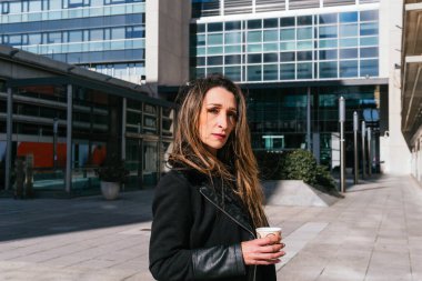 Yönetici kız kameraya bakıyor, teneffüste kahve içiyor, iş merkezinin önünde arka planda ofisler var..