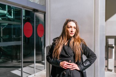 Yönetici kızın otomatikman kapısı ve arka planda yürüyen merdivenleri olan bir iş merkezinin girişinde beklediği orta boy bir fotoğraf.