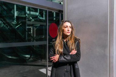 Ofis kapısında bekleyen orta ölçekli yönetici kadın. Arka planda otomatik kapı ve yürüyen merdivenler var.