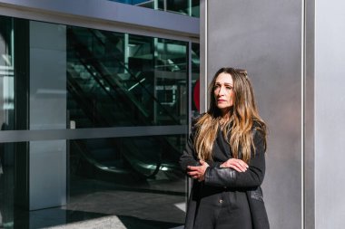 Ofis kapısında bekleyen orta ölçekli yönetici kadın. Arka planda otomatik kapı ve yürüyen merdivenler var.