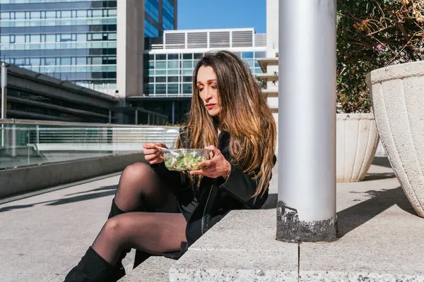 Executive girl on work break eating a plate of salad outside a business center with offices in the background