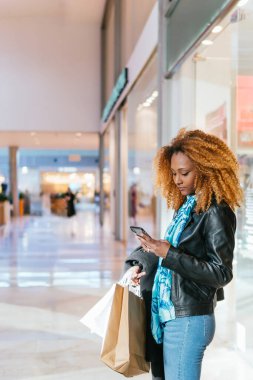 Bir alışveriş merkezinin girişinde elinde alışveriş poşetleri tutarken telefonunda afro saçlı bir kadın mesaj atıyor.