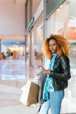 Genç siyahi bir kız bir alışveriş merkezinin girişinde elinde alışveriş torbalarıyla kameraya bakarken telefonuna mesajlar yazıyor.