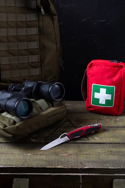 Swiss army knife and first aid kit. Binoculars and military backpack on a military box. Vertical frame. First aid kit and knife.