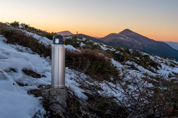 Travel thermos for hot drinks. Thermos on top of the mountain. Thermos, snow, stones and mountains. With a thermos outside. Front view.