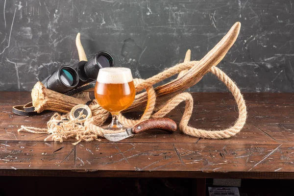 A glass of beer against the background of an elk horn. Moose horn and binoculars. Rope and binoculars. Front view.