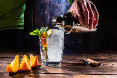 Making a cocktail. Pour the drink from a shaker. A glass with ice and mint. Orange slices and chocolate pieces on the table. Glass and ice. clipart