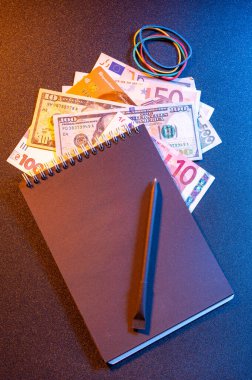 İçinde çeşitli küresel para birimleri, siyah bir defter ve karanlık bir yüzey üzerinde bir kalem bulunan sinematik finansal düzlük. Sarı ve mavi tonlarda sanatsal ışıklandırma dramatik bir atmosfer yaratır..