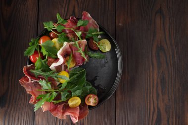 İnce dilimlenmiş jamon (tedavi edilmiş jambon) içeren gurme yemek sunumu, taze sebze ve yapraklı yeşilliklerin canlı bir karışımı ile birlikte siyah bir tabakta düzenlendi..