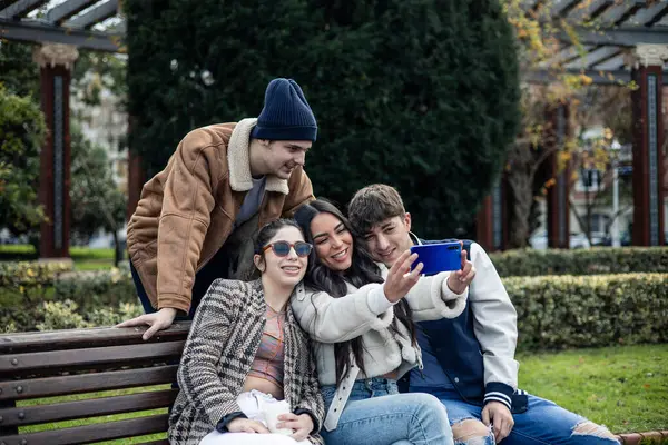 group of four young multiracial friends taking selfie with cell phone sitting on a park bench