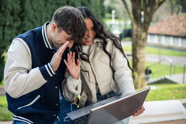 couple of latin girl and caucasian boy waving at laptop screen with headphones making video call outdoors