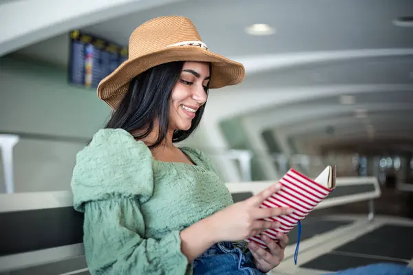 closeup young latina with hat reading a book sitting in the station terminal while waiting