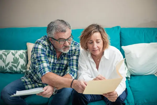 a retired couple sits comfortably on their sofa, diligently sorting through papers and documents. One of them wears glasses, symbolizing focused attention to detail. This image captures the essence of partnership in financial management during retire