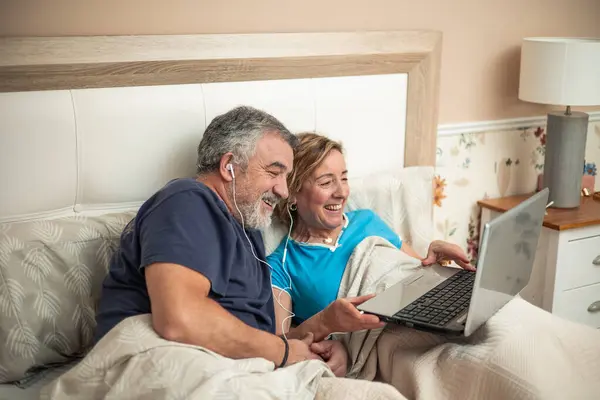 Two retired couple relax in the comfort of their home, lying on the bed with a laptop and headphones. They are seen smiling and laughing as they enjoy online activities together, demonstrating that retirement can be a time of connection,