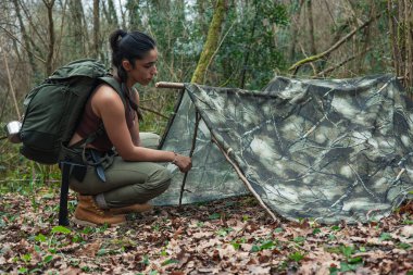 Image of a young adventurer woman crouching down in front of the self-built shelter she has constructed in the forest, showcasing her outdoor survival skills clipart
