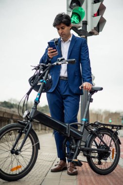 Dikey portre. Takım elbiseli bir işadamı elektrikli bisiklet ve kaskını tutarken akıllı telefonuyla mesaj gönderiyor. Modern şehir yaşam tarzında sürdürülebilir hareketlilik