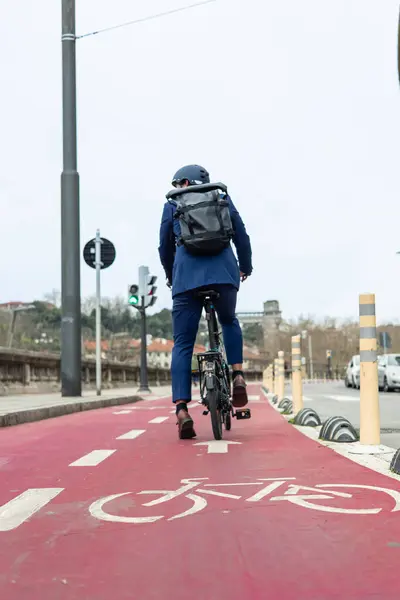 A male businessman, seen from behind, is commuting with a backpack on his electric folding bike along a red bike lane in the city. He is on his way to work