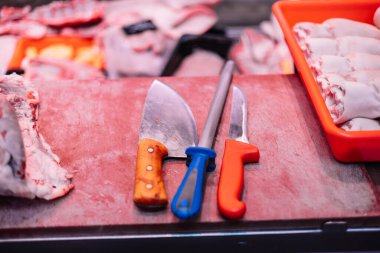 Kasap dükkanındaki et kesme tahtasına yerleştirilmiş iki kasap bıçağı ve bir bıçak bileyicisi. Kasap sanatı için gerekli aletler et hazırlama konusunda gerekli hassasiyet ve uzmanlığı yansıtıyor.