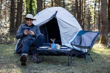 Neşeli bir kampçı, kaşif şapkası takıyor, çadırının önünde oturuyor, akıllı bir telefonla sohbet ediyor ve yaz günbatımından önce doğanın ortasında bir kamp masası kuruluyor.