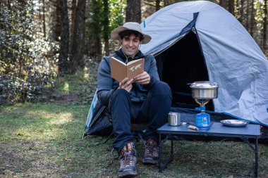 Maceraperest bir adam, kaşif şapkası takıyor orman kamp alanında kitap okuyor, çadırı önünde oturuyor ve yemeği kamp ocağında pişiyor.