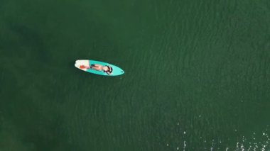 İHA antenli bikinili Latin kadın yaz günü güneşlenirken rahatça sörf tahtasına uzanıyor.