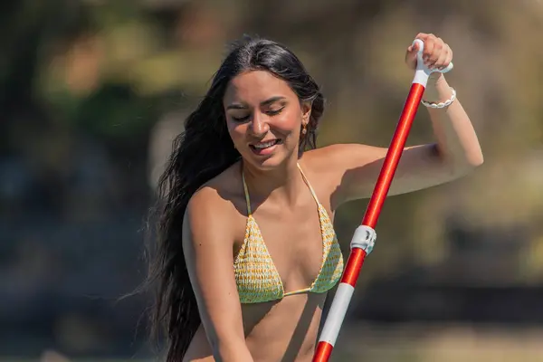 closeup smiling multicultural Latina woman in a bikini is paddling gracefully on her paddleboard on a sunny summer day at the beach. Her cheerful expression setting convey a sense of joy and relaxation