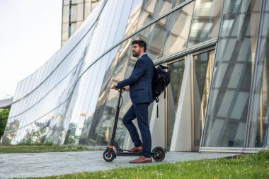 Takım elbiseli ve sırt çantası taşıyan iş adamı bir finans binasının önünde elektrikli scooterının yanında duruyor. Kurumsal dünyada modern ve sürdürülebilir işe gidip gelme uygulamalarını örneklendiriyor.