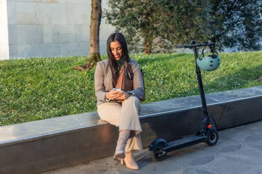 İş kadını, elektrikli scooterının yanındaki bankta oturan finans bölgesinin koşuşturmasına kısa bir süre ara veriyor. Akıllı telefonuna dalmış, bir mesaj yazıyor.