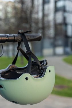 Elektrikli scooter 'ın gidonuna asılı dikey yakın plan güvenlik kaskı, şehir içi ulaşımı sembolize ediyor. Görüntü, modern hareketliliğin ve güvenlik bilincinin özünü yakalıyor.