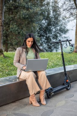 Dikey iş kadını elektrikli scooterının yanındaki bankta oturuyor ve dizüstü bilgisayarına odaklanıyor. Mobilite ve teknolojinin modern karışımını vurguluyor, esnek bir çalışma ortamı sergiliyor