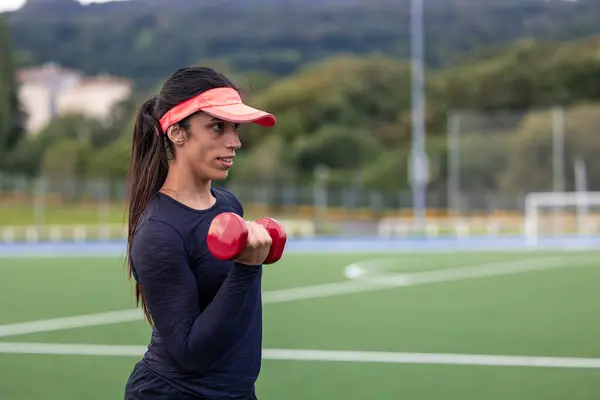 Kapalı alan bayan sporcusu, antreman sırasında kollarını ve kollarını çalıştırmak için bir dambılı kaldırıyor. Görüntü, açık hava ortamında dayanıklılık eğitimi ve uygunluğa olan bağlılığını vurguluyor.