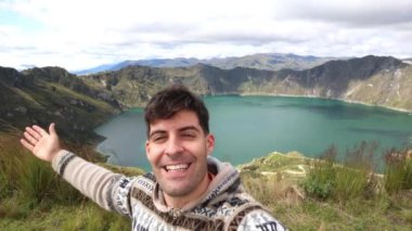 Gülümseyen Latin turist, Ekvador 'daki Quilotoa Volkanı' ndaki (3,914 metre) lagünün canlı mavi sularını gösterirken selfie çekiyor. Bu neşeli an And manzarasının güzelliğini yakalar.