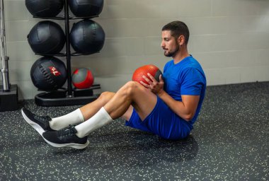 Genç sporcu spor salonunda oturmuş bir ilaç topuyla çekirdek egzersizleri yapıyor. Bu çalışma çekirdek kuvveti ve istikrarına odaklanır, fitness ve atletik gelişime olan bağlılığını gösterir.